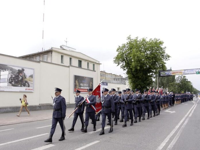 Funkcjonariusze zakładu karnego, w mundurach, idą środkiem ulicy trzymając sztandar