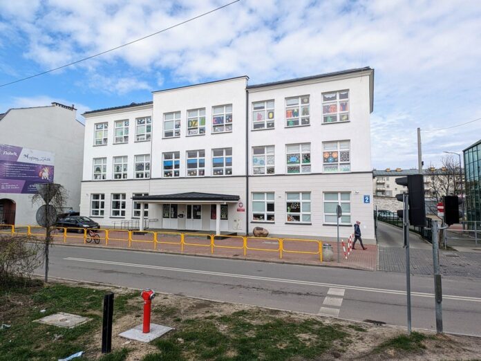 Na zdjęciu widok na front jasnego trzykondygnacyjnego budynku szkoły z kolorowymi naklejkami na szyby.