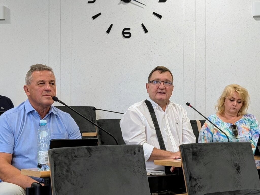 Dwaj mężczyźni i kobieta siedzą na krzesłach, przed nimi mikrofony