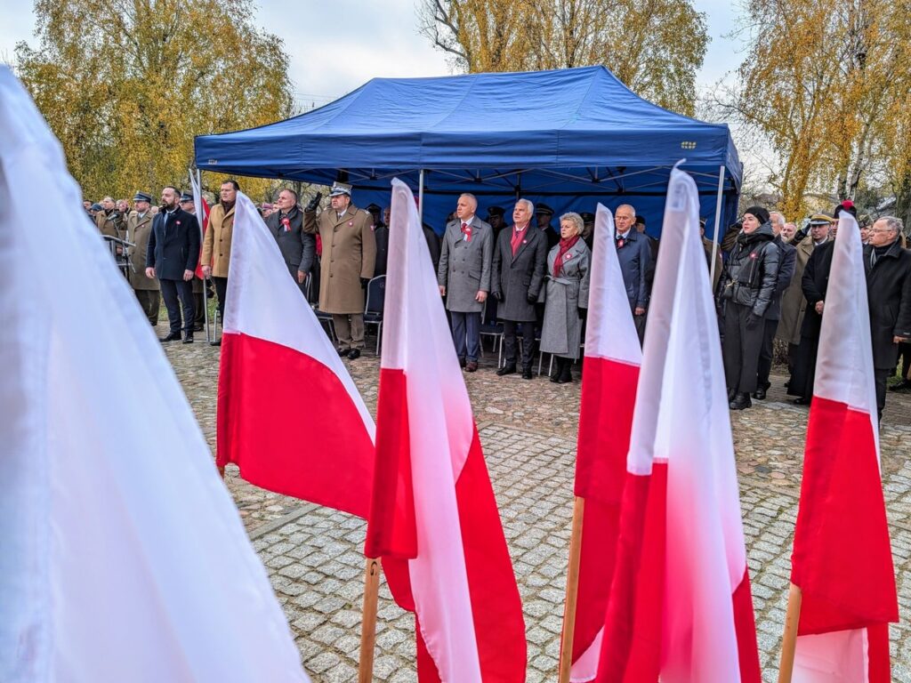 Grupa ludzi stoi wokół biało-czerwonej flagi, symbolizując jedność i patriotyzm. 