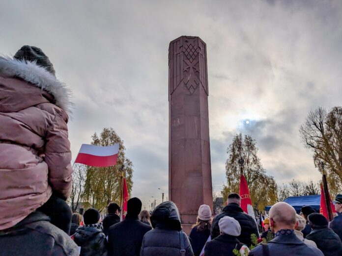 Grupa osób stoi zwrócona twarzą w kierunku wysokiego pomnika.
