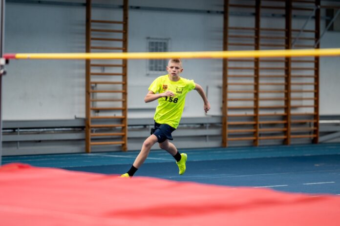 Młody mężczyzna biegnie po torze w hali sportowej, skupiony na osiągnięciu jak najlepszego wyniku.