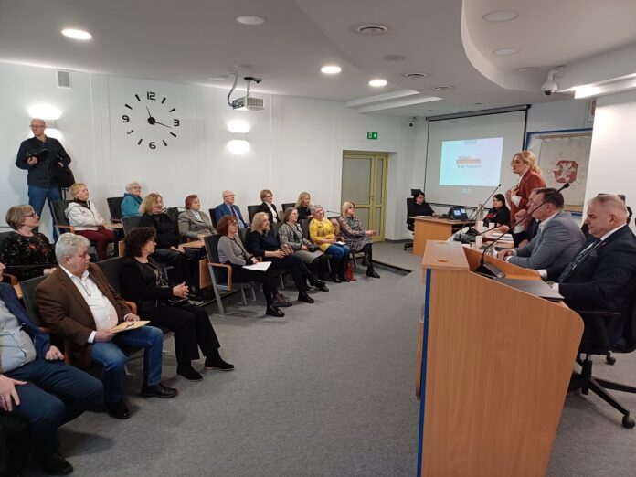 Grupa ludzi siedzi w sali, jedna z osób przemawia z podium, prowadząc dyskusję lub prezentację.