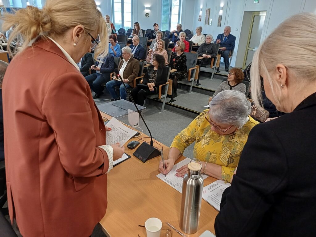 Duża grupa ludzi siedzących na krzesłach w sali, zróżnicowane postawy i wyrazy twarzy uczestników.