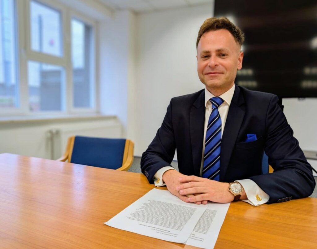 Mężczyzna w garniturze siedzi przy stole, przeglądając dokumenty leżące przed nim.