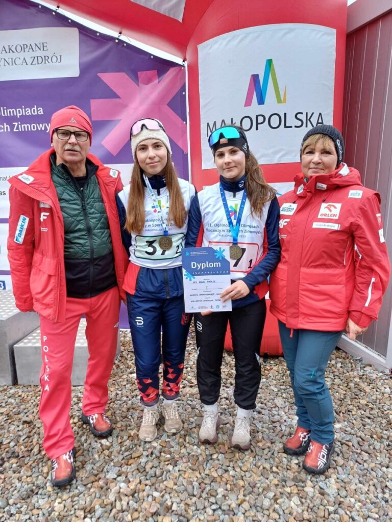 Trzy osoby w czerwonych i niebieskich kurtkach trzymają medal, uśmiechając się do kamery.