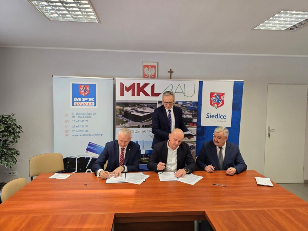 Trzech mężczyzn siedzących przy stole, podpisujących dokumenty w formalnej atmosferze.