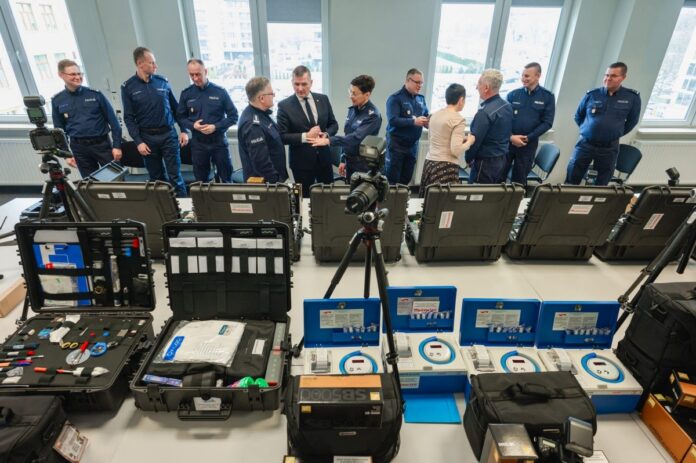 Grupa ludzi stoi wokół stołu z różnorodnym sprzętem, angażując się w dyskusję i współpracę.