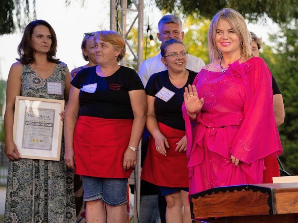 Kobieta w różowej sukience na tle grupy osób, uśmiecha się i macha do publiczności. 