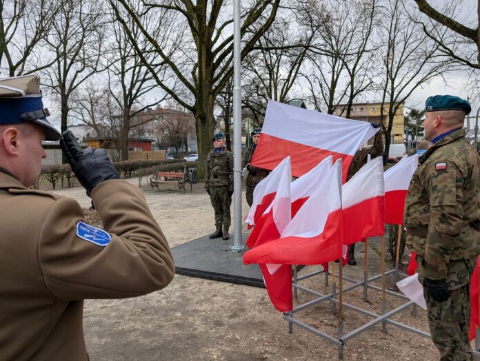 Grupa żołnierzy w mundurach stojących przy powiewających flagach Polski podczas uroczystości