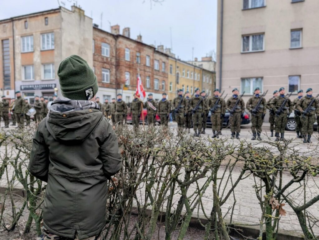 dziecko stoi w kurtce i czapce i patrzy na stojących w rzędzie żołnierzy w mundurze prezentujących broń.