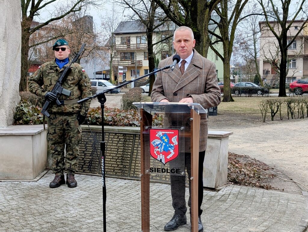Mężczyzna w garniturze stoi przy mikrofonie, w tle żołnierz w mundurze trzyma broń