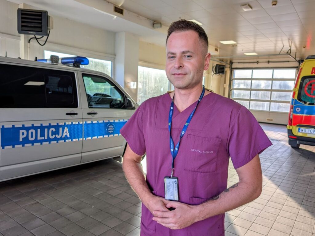 Mężczyzna z lekarskim stroju stoi na tle radiowozu policyjnego i karetki pogotowia. 