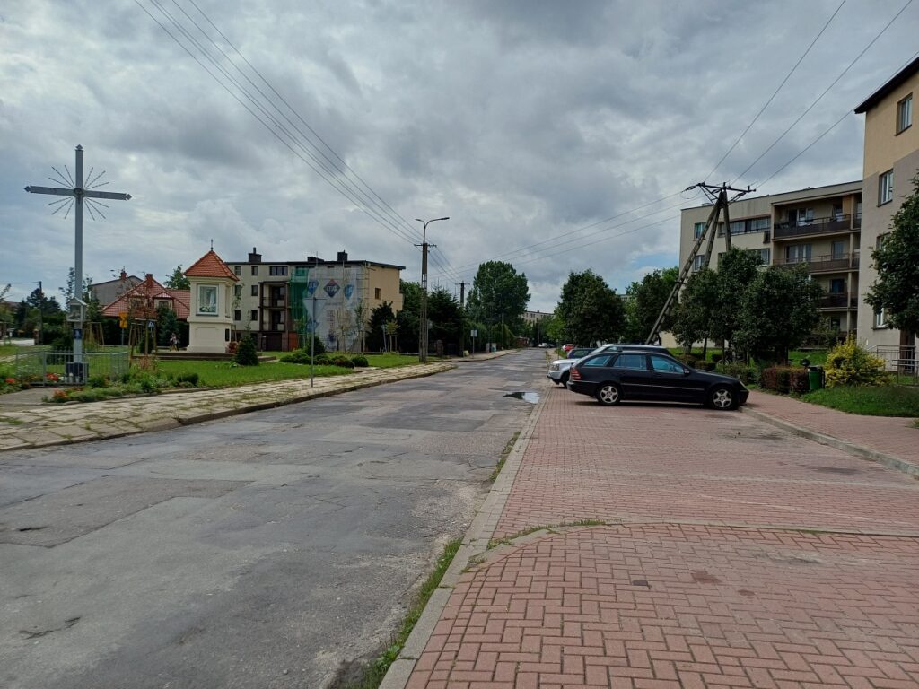 Ulica ze zniszczoną nawierzchnią, z chodnikiem i zaparkowanymi samochodami. 