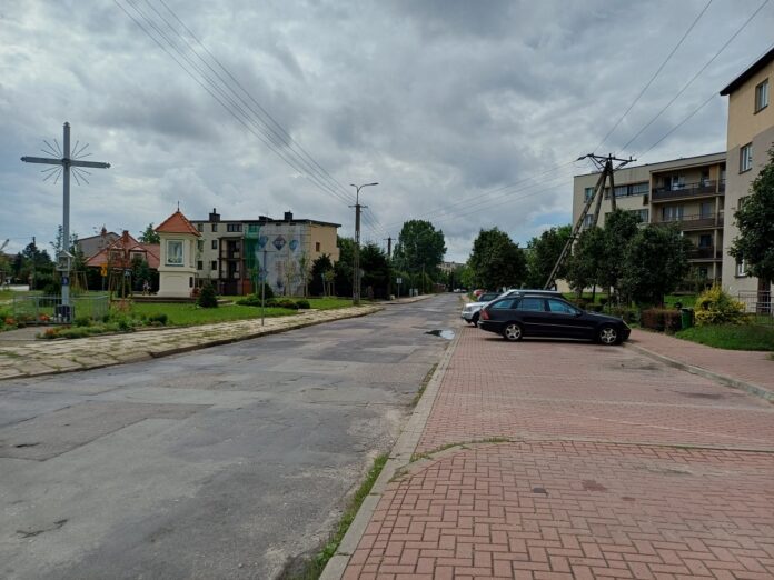 Ulica ze zniszczoną nawierzchnią, z chodnikiem i zaparkowanymi samochodami.
