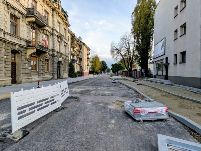 Miejska ulica w budowie, widać nowy asfalt, po obu stronach zabudowania.
