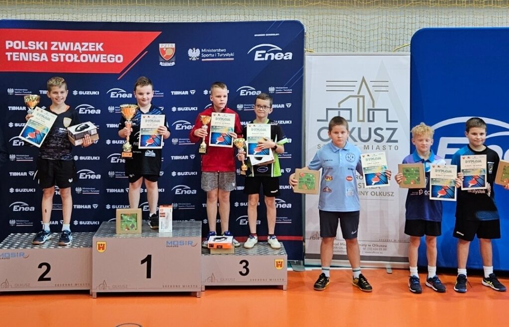 Grupa chłopców na podium trzymających swoje trofea, uśmiechniętych i radosnych po zwycięstwie.