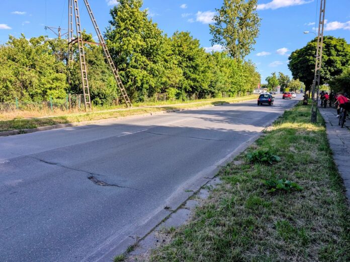 Zniszczona asfaltowa ulica dwukierunkowa w mieście otoczona zielenią.