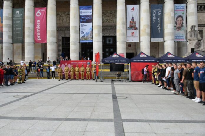 Grupa kilkunastu osób w tym strażaków stojąca przed budynkiem z kolumnami.