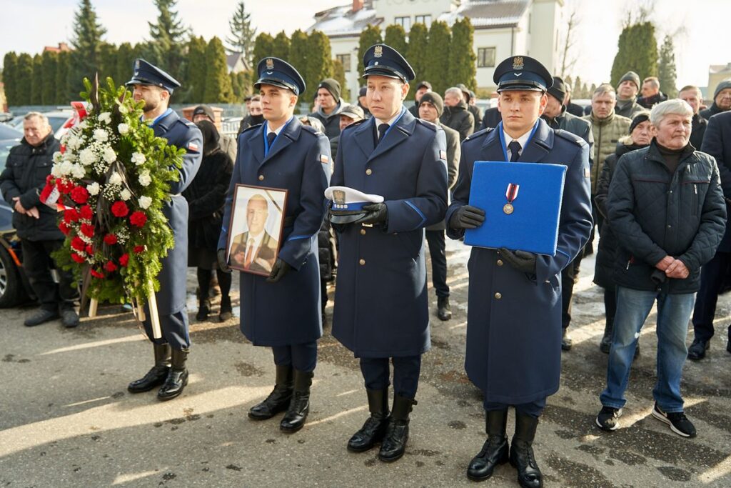 Czterech policjantów w galowych strojach bierze udział w uroczystościach pogrzebowych swojego kolegi. Trzymają zdjęcie, wiązankę, order i czapkę.