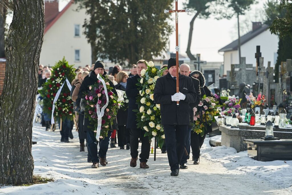 Uroczystości pogrzebowe grupa osób niesie wiązanki i krzyż