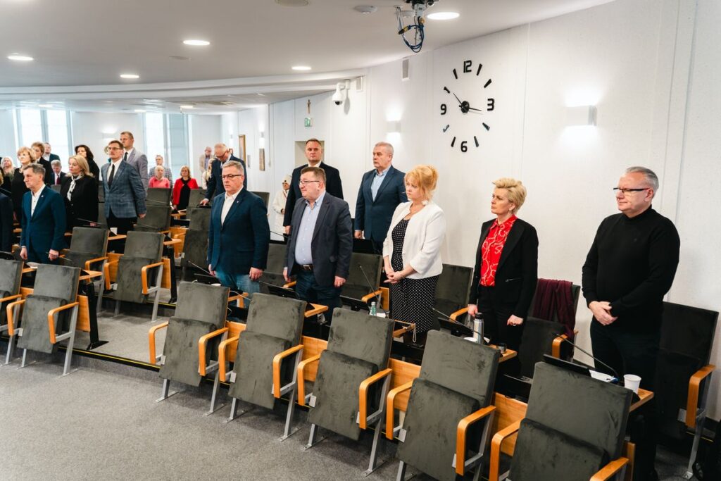 Grupa mężczyzn i kobiet stoi w skupieniu i zadumie w sali konferencyjnej urzędu miasta. 