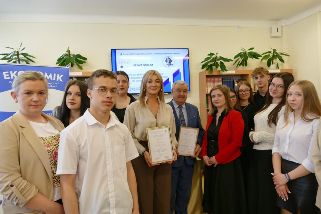 Grupa ludzi stoi przed telewizorem, zwracając uwagę na wyświetlany program.