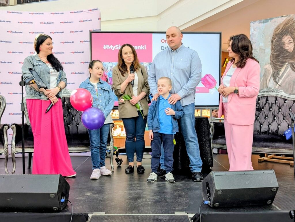 Rodzina stoi na scenie z balonami, a kobieta trzyma mikrofon i przemawia do publiczności.