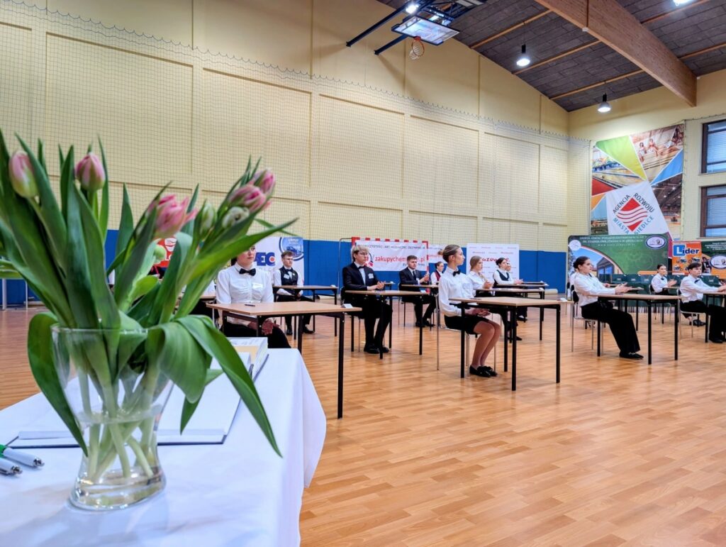 Grupa ludzi siedzi przy stołach w gimnazjum, rozmawiając i spędzając czas razem.