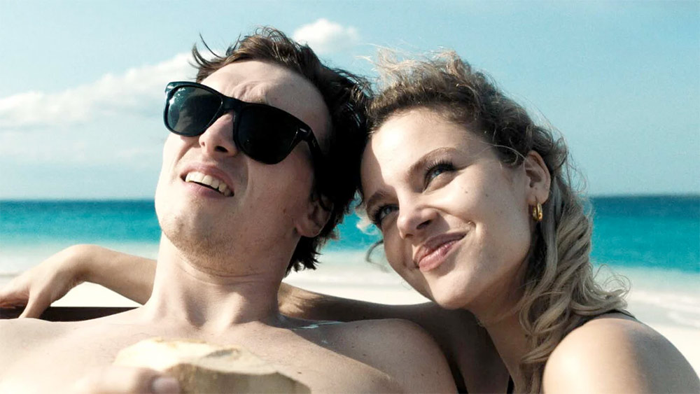 Mężczyzna i kobieta siedzą na plaży, trzymając kokos, otoczeni piaskiem i falami morza.