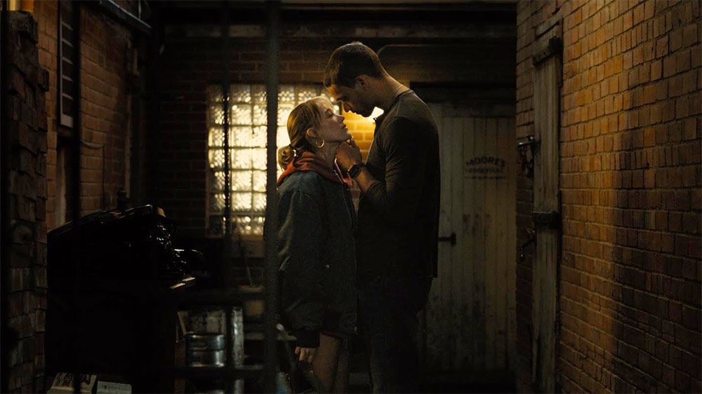 Mężczyzna i kobieta stoją w wąskiej alejce, otoczeni murami i delikatnym światłem.