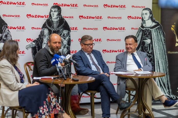 Trzech mężczyzn i jedna kobieta siedzą przy stole z mikrofonami, prowadząc dyskusję.