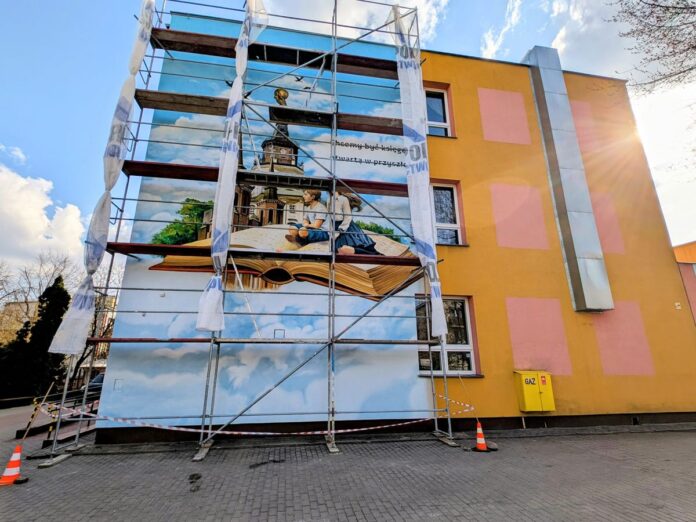 Mural na budynku przedstawiający chłopca i dziewczynkę siedzących na otwartej książce.