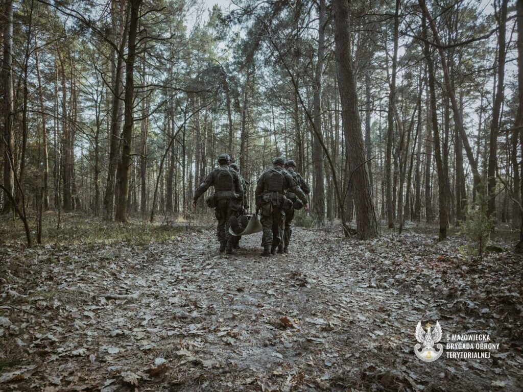 Dwaj żołnierze idą przez las, niosąc nosze.