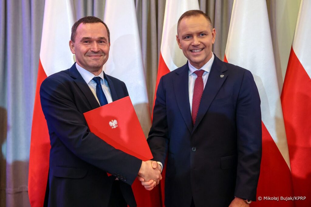 Dwaj uśmiechnięci mężczyźni, jeden z nich trzyma w dłoniach książkę z czerwonymi okładkami.