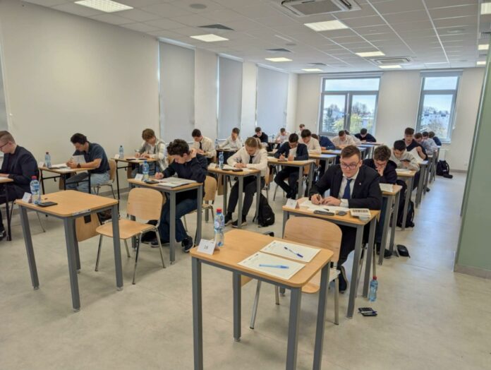 Wnętrze klasy z uczniami siedzącymi przy biurkach, zaangażowanymi w naukę.