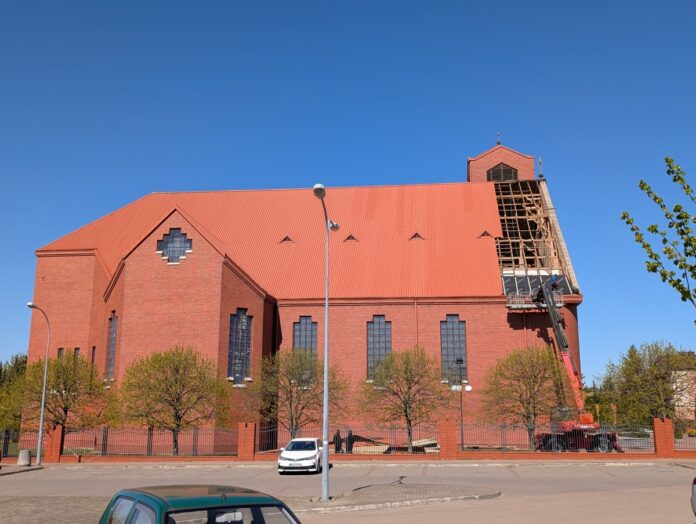Czerwony kościół z cegły z dużym dachem, otoczony zielenią, w słoneczny dzień.