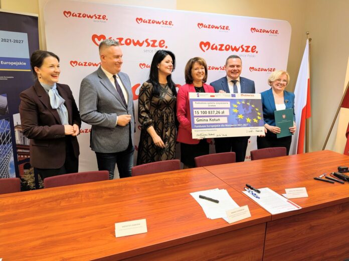 Grupa uśmiechniętych i zadowolonych osób trzyma w ręku duży karton z informacją o dotacji unijnej.