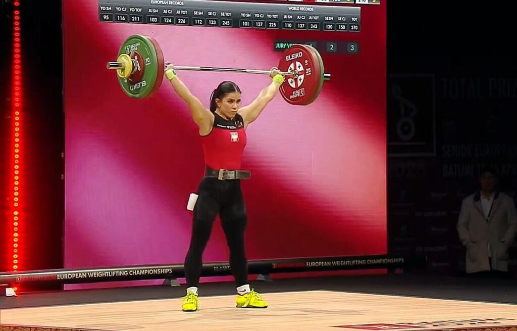 Kobieta podnosi ciężar na scenie, prezentując siłę i determinację.