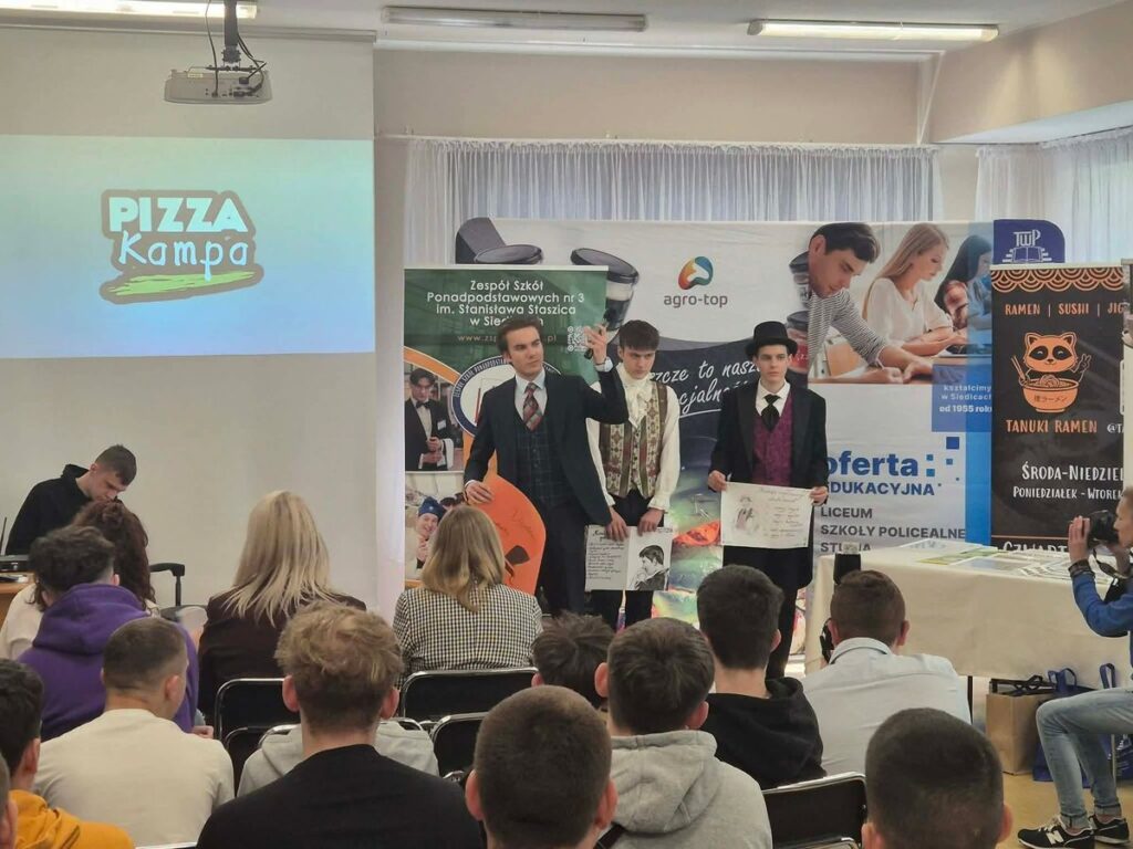 Grupa zwycięzców w radosnej atmosferze trzyma puchary, w tle widać innych uczestników.