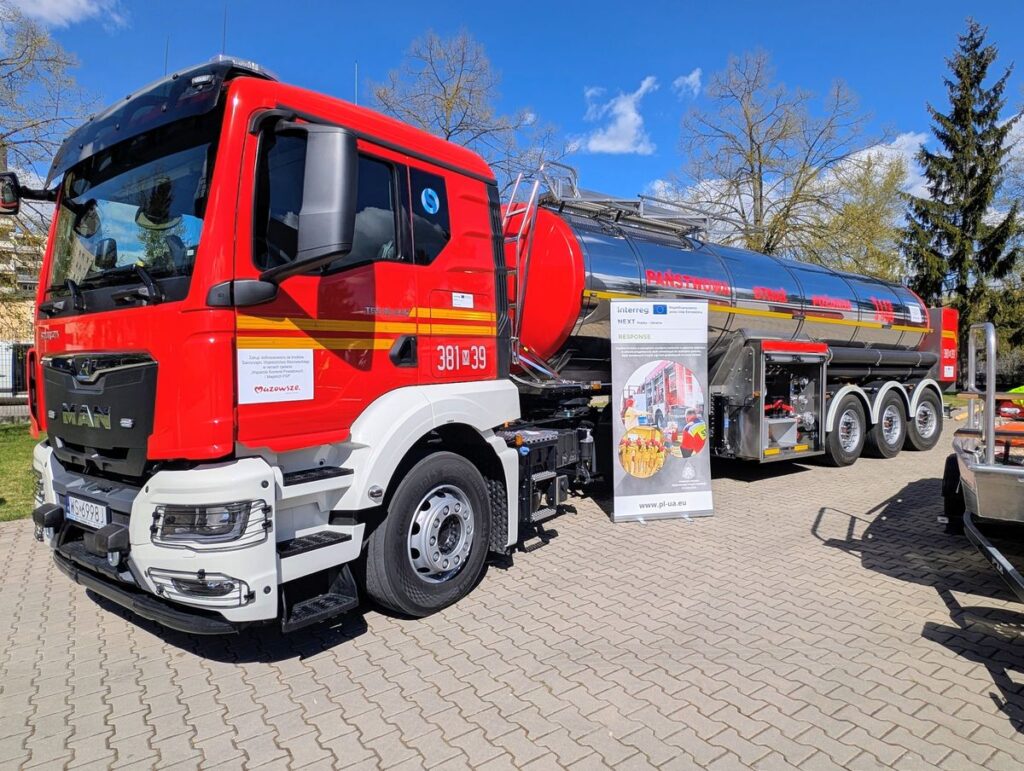 Duży czerwono-biały samochód cysternowy zaparkowany przed budynkiem.