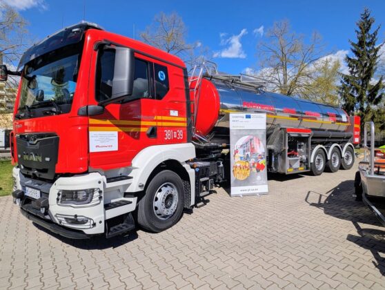 Duży czerwono-biały samoch&oacute;d cysternowy zaparkowany przed budynkiem.