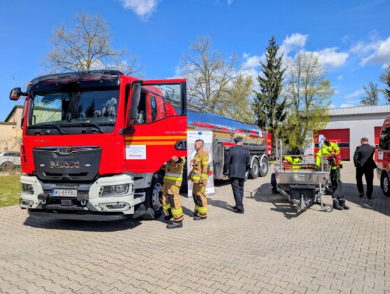 Duży czerwono-biały samoch&oacute;d cysternowy zaparkowany przed budynkiem oraz stojący strażacy