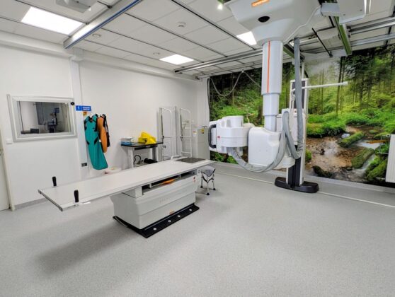 Szpitalny pok&oacute;j z dużym sprzętem medycznym, w tle widoczne ł&oacute;żko pacjenta i okno z naturalnym światłem.