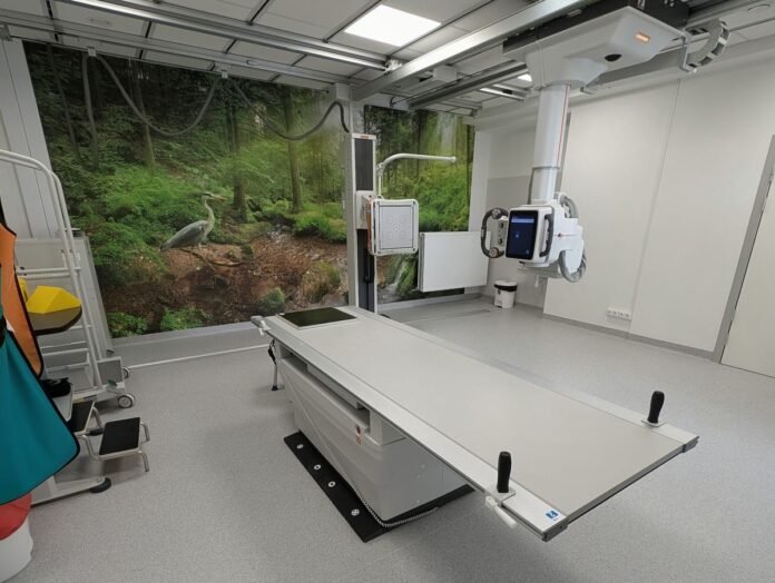 Szpitalny pokój z dużym stołem medycznym, otoczony sprzętem medycznym i neutralnymi ścianami.