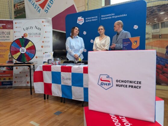 Trzy osoby stoją obok stołu z widocznym znakiem na nim.