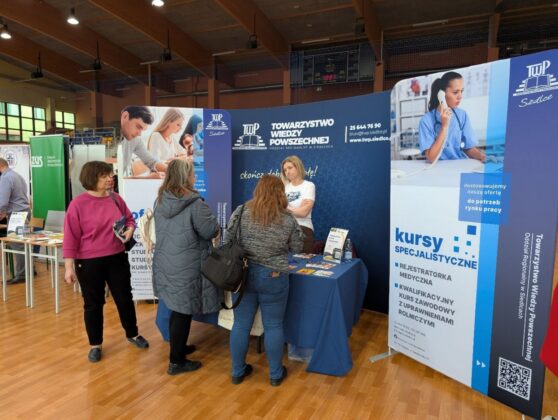 Ludzie stoją wok&oacute;ł stołu z banerem i znakiem, uczestnicząc w wydarzeniu lub spotkaniu