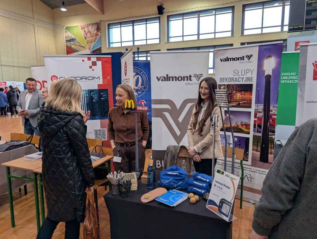 Ludzie stoją wokół stołu z różnymi przedmiotami, rozmawiając i przeglądając zgromadzone rzeczy.