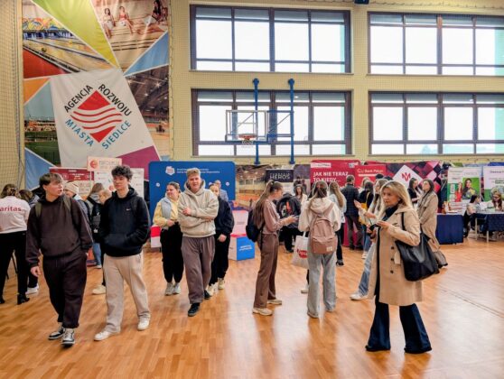 Ludzie stoją w dużym pomieszczeniu, otoczeni licznymi plakatami na ścianach.