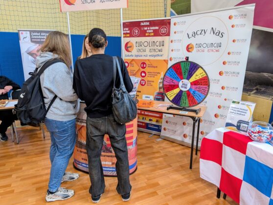 Dwoje ludzi stoi przy stole z kołem i znakiem, rozmawiając i wskazując na przedmioty na stole.